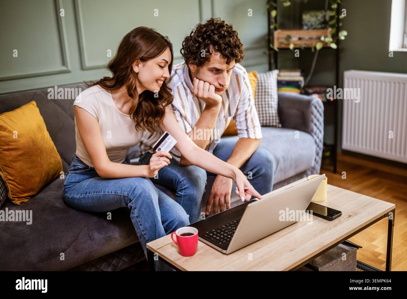 A young couple sits together on a sofa, comparing options on a laptop while making an online purchase. The woman holds a credit card and points at the Stock Photo