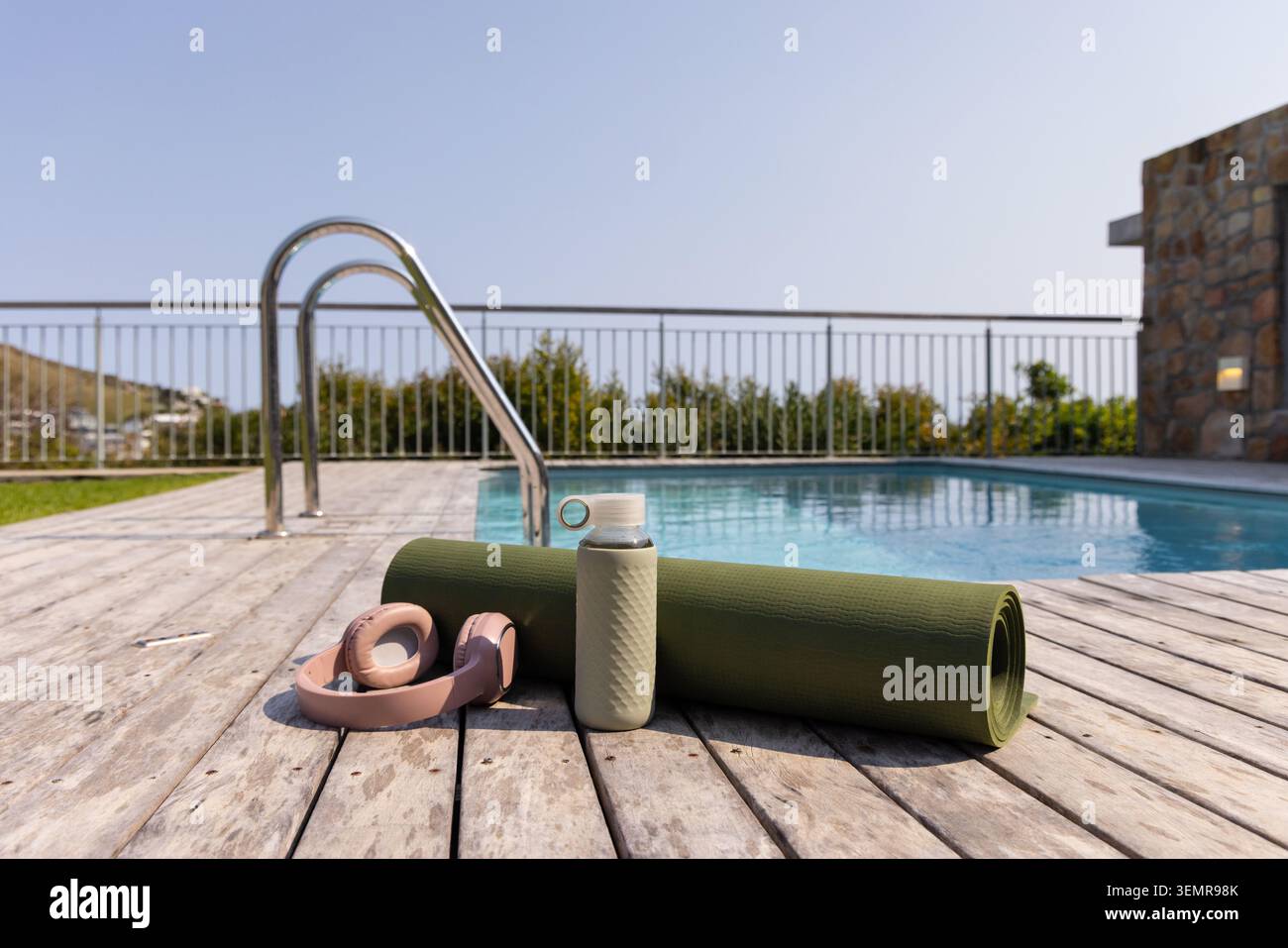 Rolled green yoga mat with textured bottle and pink headphones resting on wooden deck by pool Stock Photo
