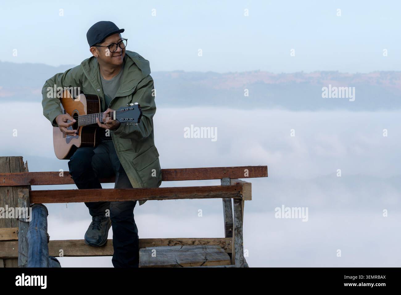 A young tourist plays an acoustic guitar on a wooden balcony at a mountain summit during the morning. Stock Photo