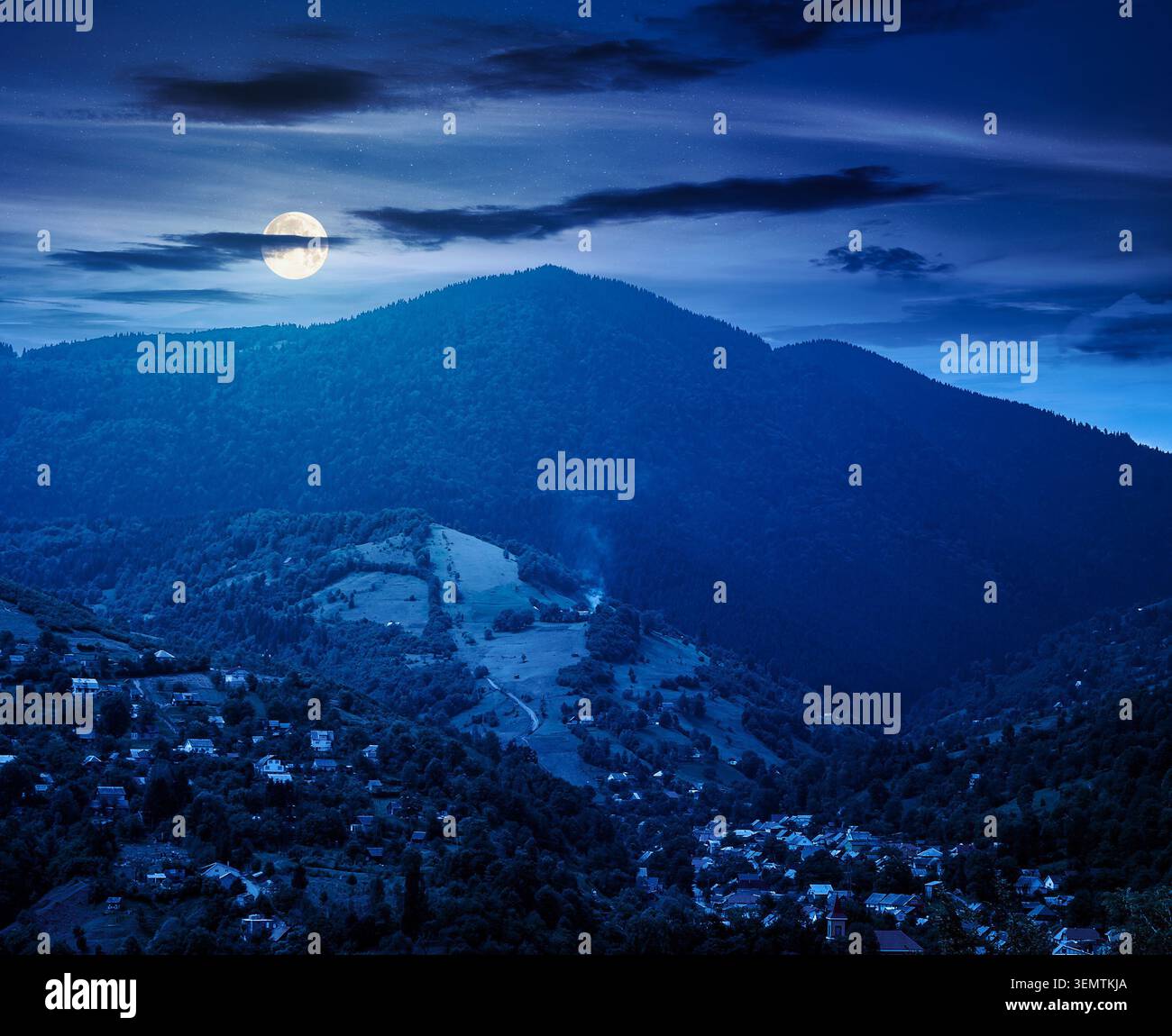 carpathian mountain landscape on summer night. full moon over view in to the valley of town near forested hill of mountain range. mysterious travel ba Stock Photo