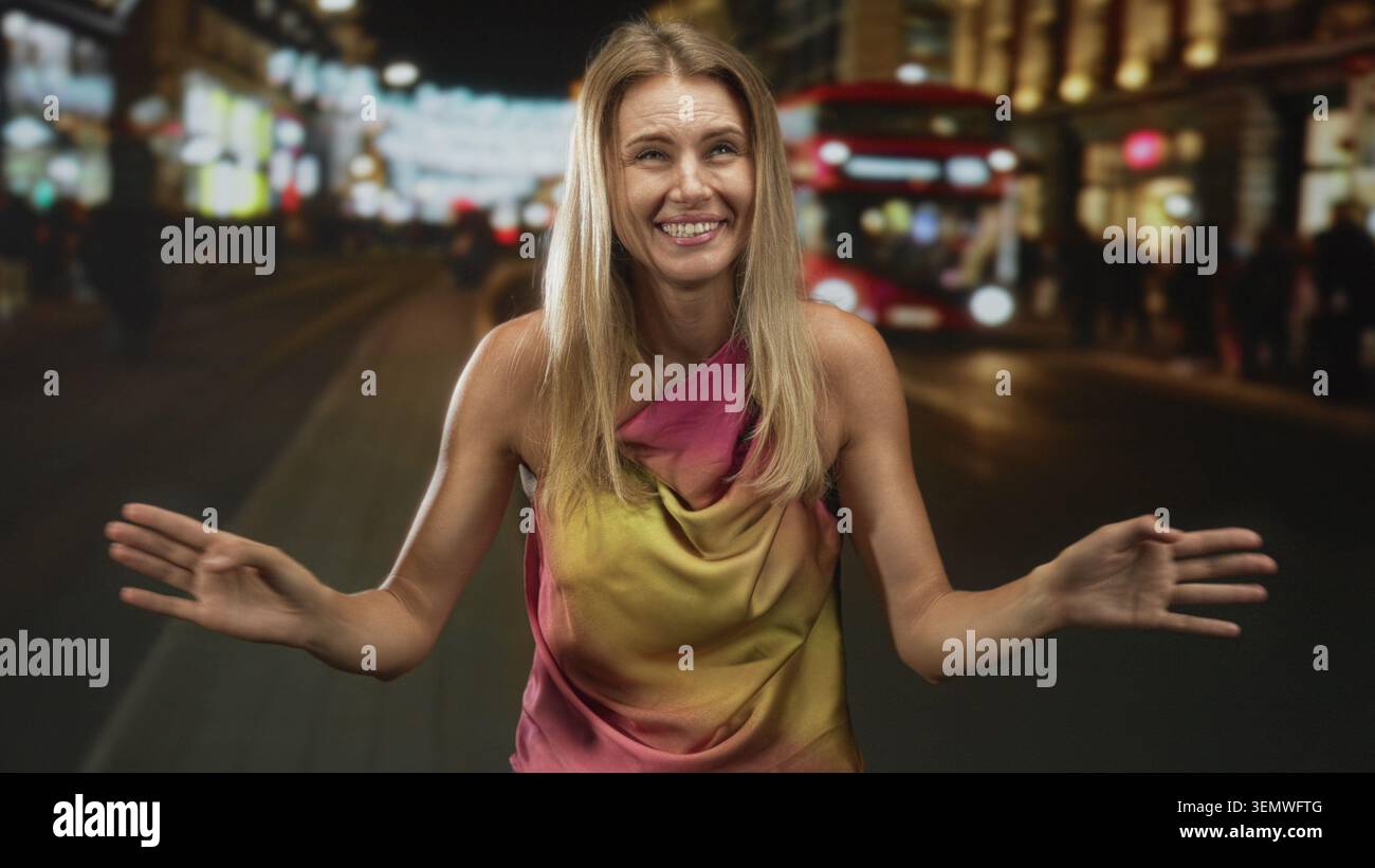 Blonde woman smiling and dancing with raised hands near a red double decker bus in a night city street; joyful. Stock Photo
