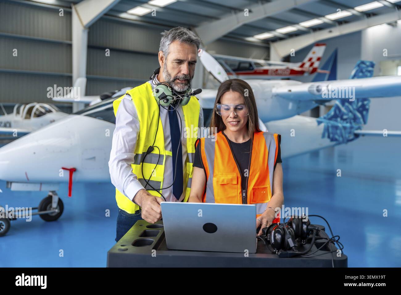 Two aviation engineers in high visibility vests and safety glasses review laptop data and discuss maintenance and inspection plans inside a hangar wit Stock Photo