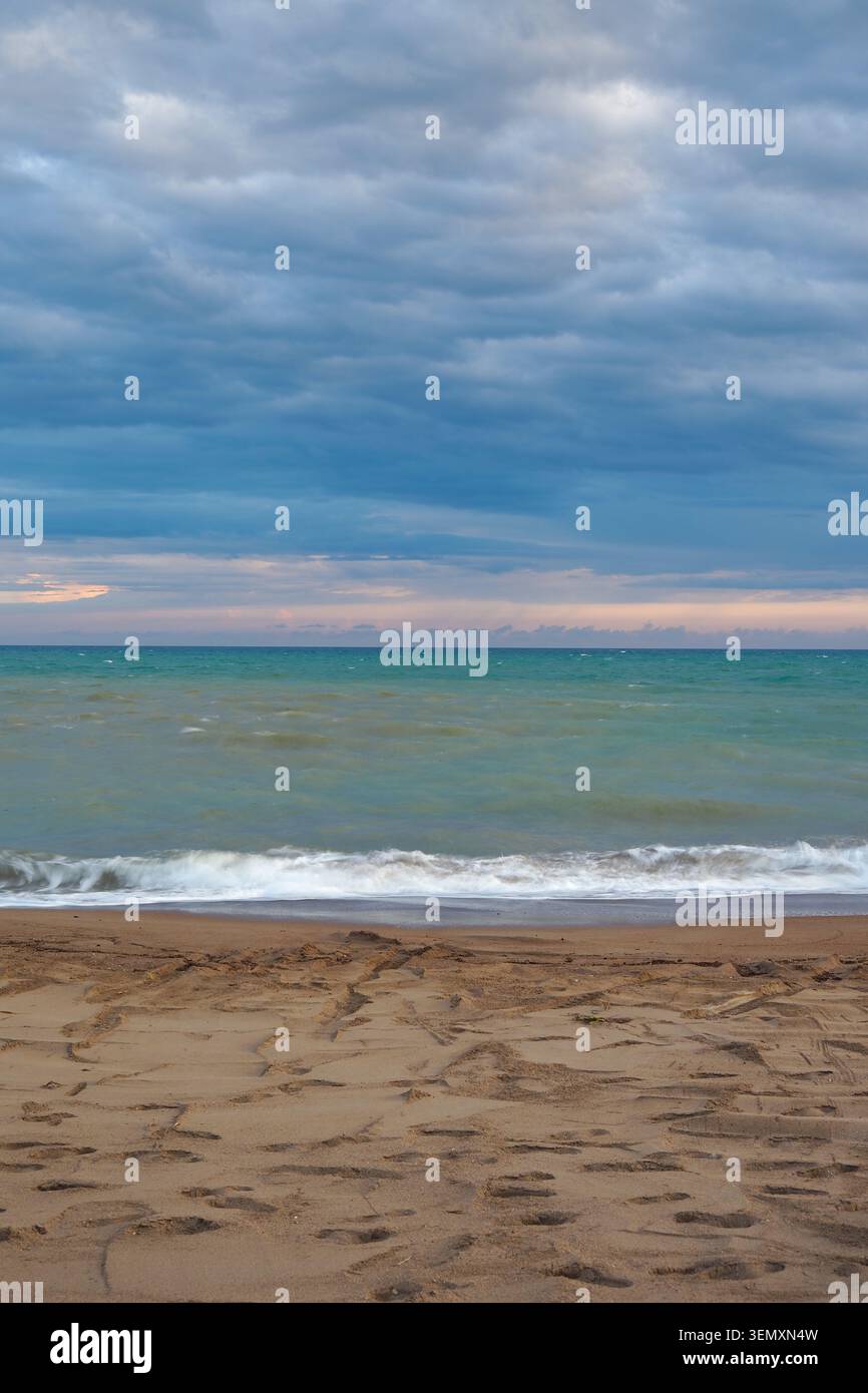 Waves crash on sandy beach under cloudy sky at sunset Stock Photo