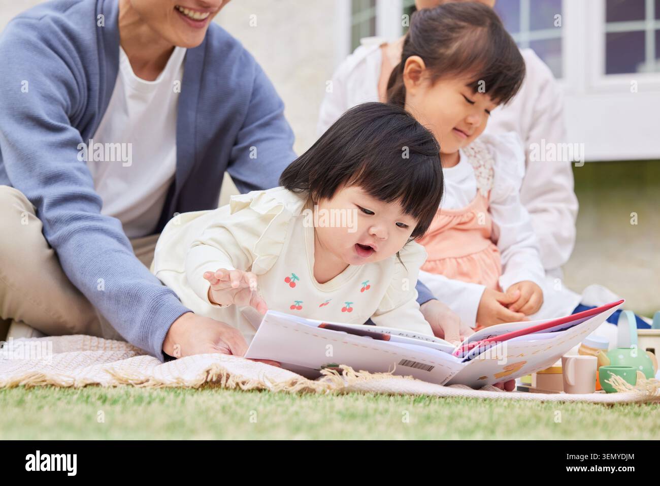Japanese family parents and two daughters an infant and a preschooler reading a picture book on a blanket outdoors Stock Photo