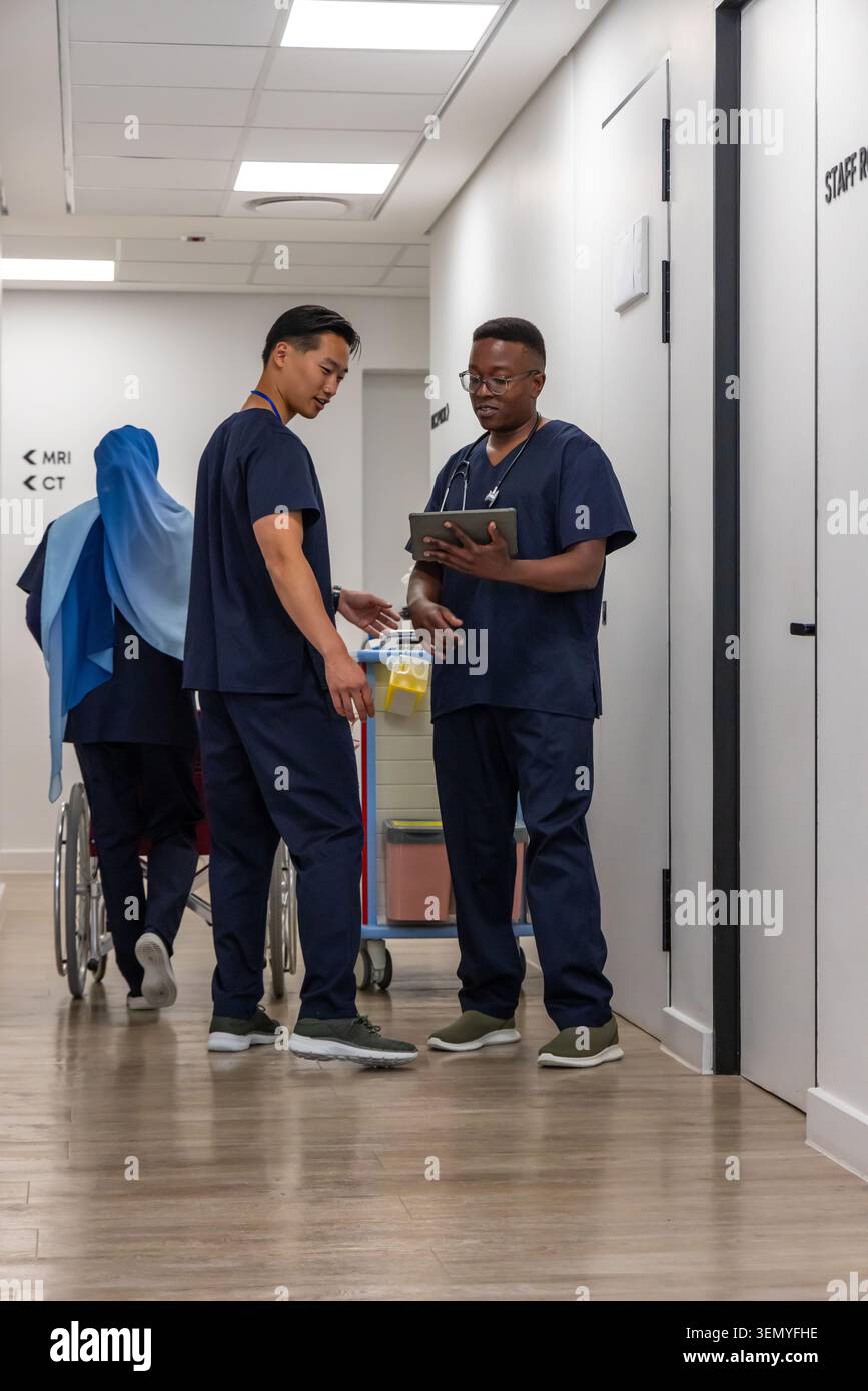 Diverse medical staff in navy scrubs consulting tablet and pushing wheelchair in hospital corridor Stock Photo
