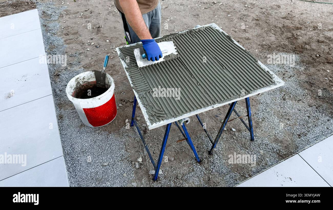 Construction worker applying adhesive mortar to a tile surface using a trowel on a work table outdoors Stock Photo