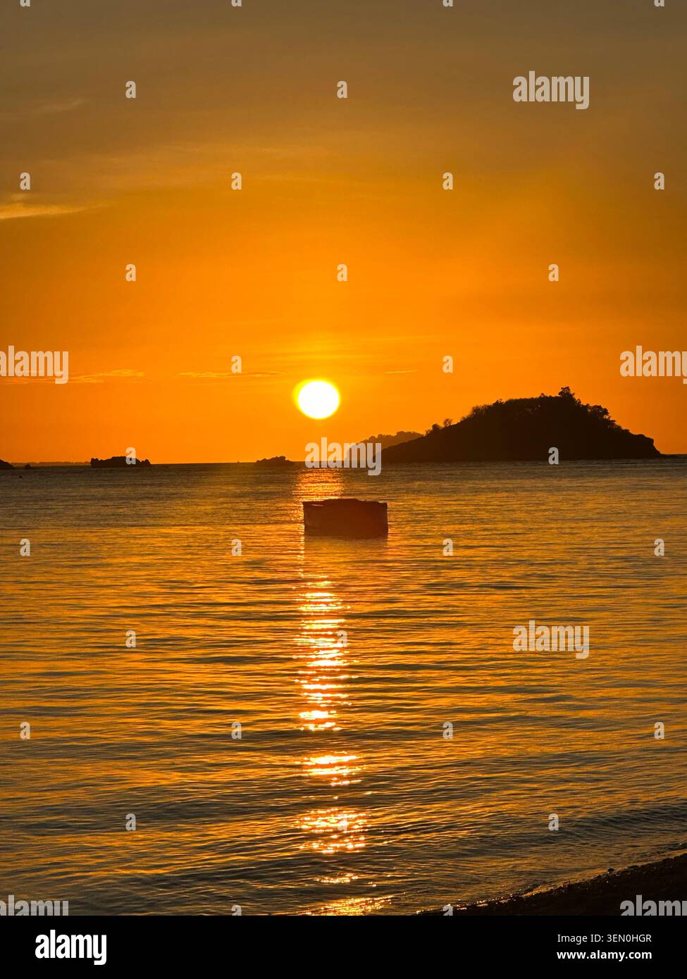 A golden sun sinks beside a dark island, drawing a molten ribbon of light across the calm evening sea - Smartphone Captured Stock Image