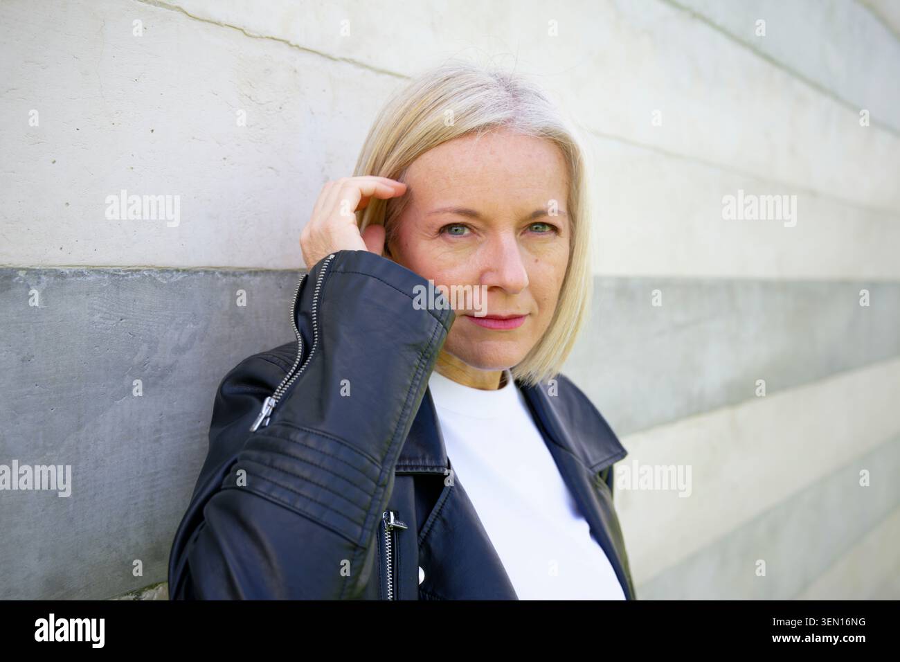 Mature woman in black leather jacket leaning against a modern concrete wall. Stock Photo
