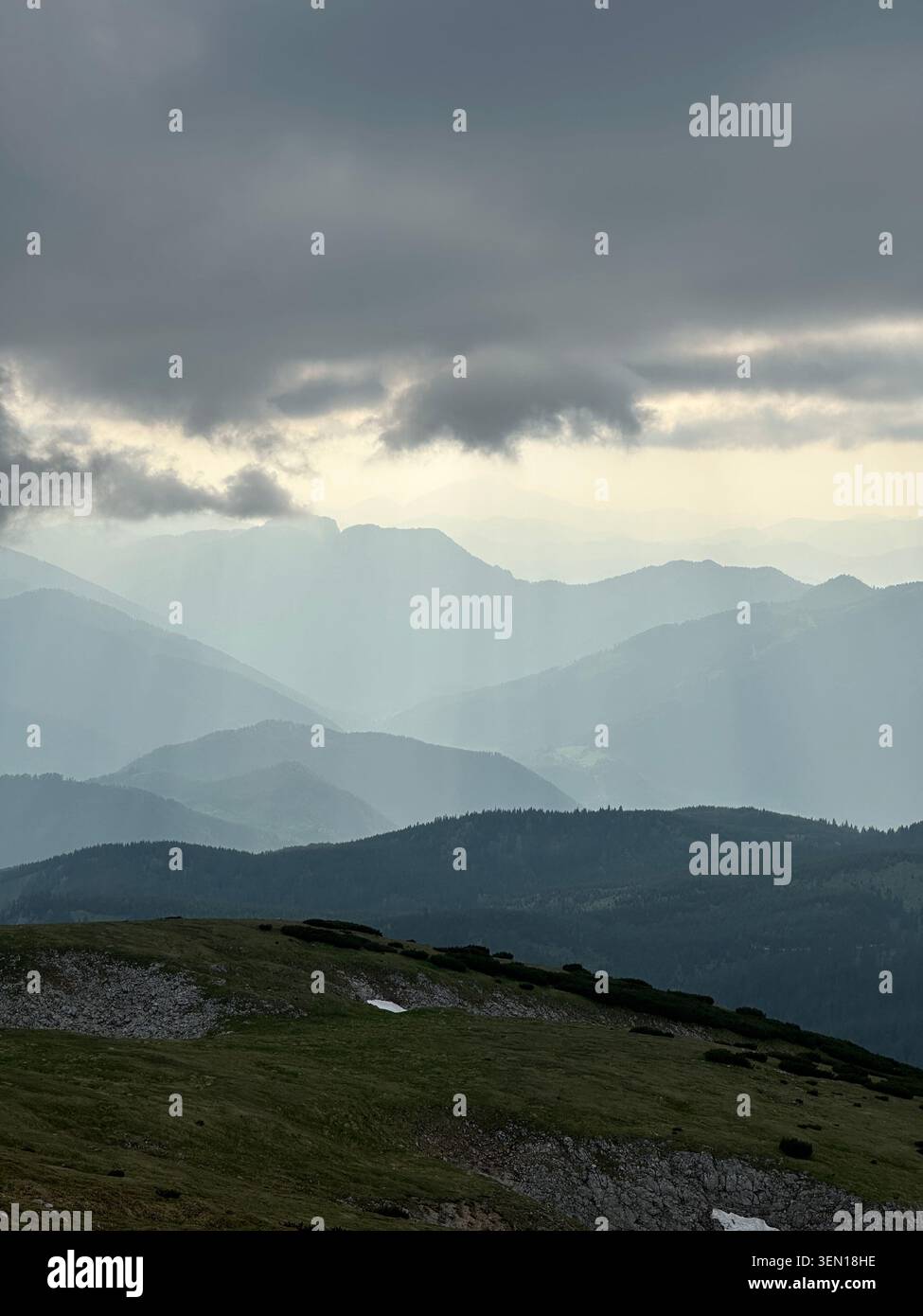 Dark storm clouds press over layered mountain ridges while pale shafts of rain and light sweep across the distant valley. 3 Stock Photo