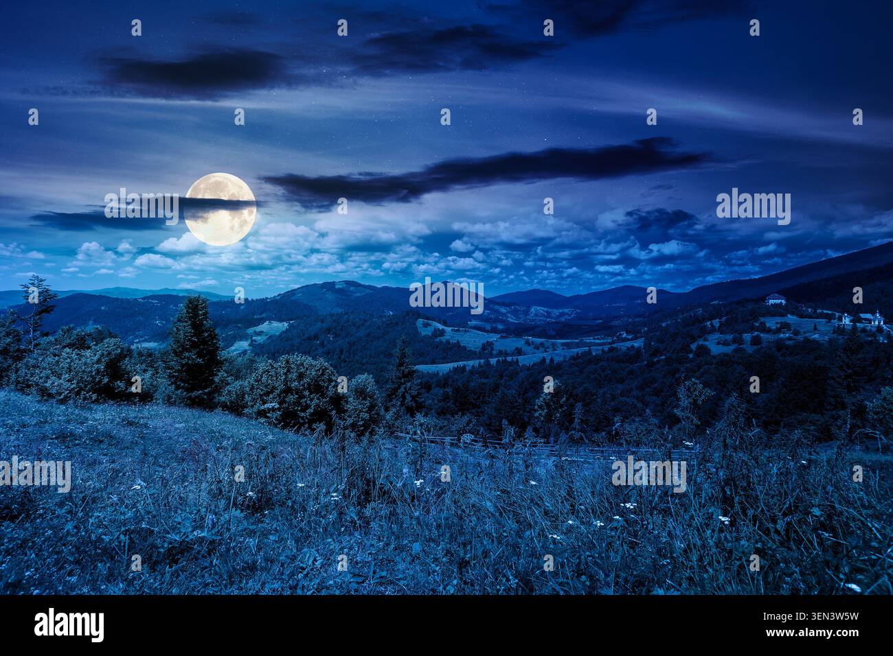 mysterious field on hillside in mountains near village at night. full moon over carpathian mountain landscape in summer. countryside view of alpine fi Stock Photo