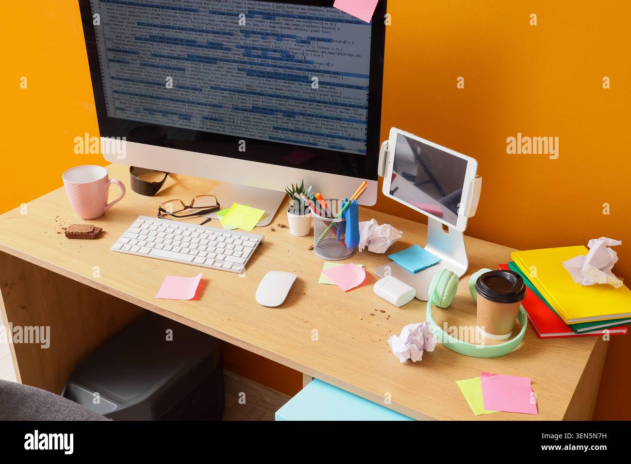 Messy programmer's desk with computer screen displaying code, tablet, coffee, and snacks on wooden table Stock Photo