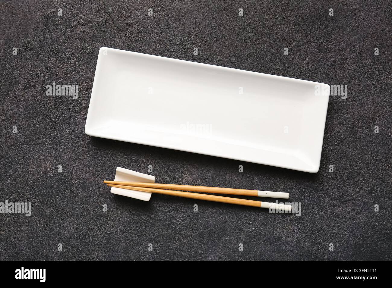 Empty white rectangular ceramic plate with wooden chopsticks on dark textured background, top view for food presentation Stock Photo