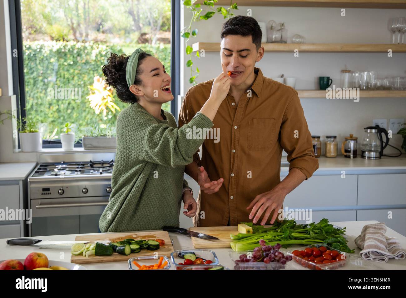 Couple laughing and feeding while preparing cucumbers, cherry tomatoes, knives on kitchen island Stock Photo