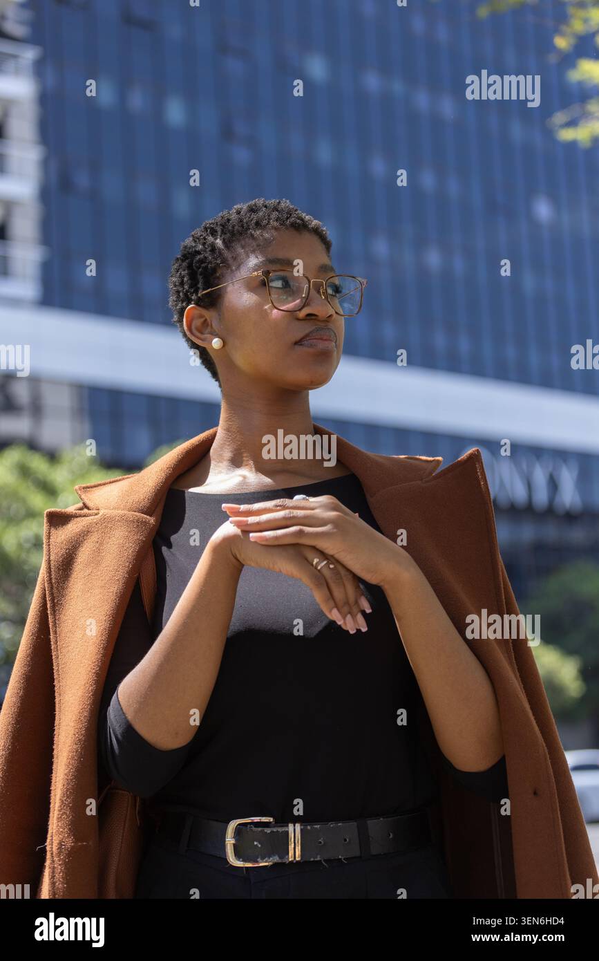 African woman standing in front of glass office building, brown coat draped, hands clasped, glasses Stock Photo