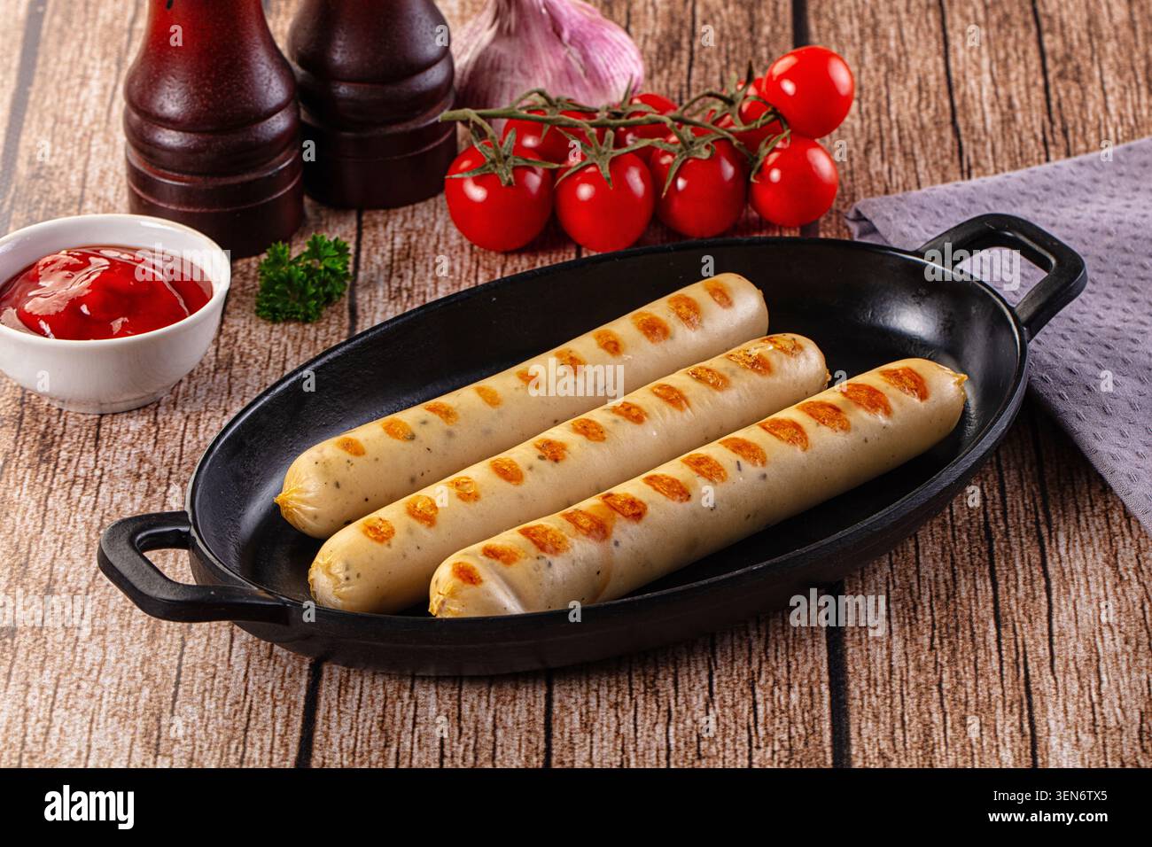 Three golden grilled sausages served in black cast iron skillet on rustic wooden table with cherry tomatoes and ketchup side dish Stock Photo