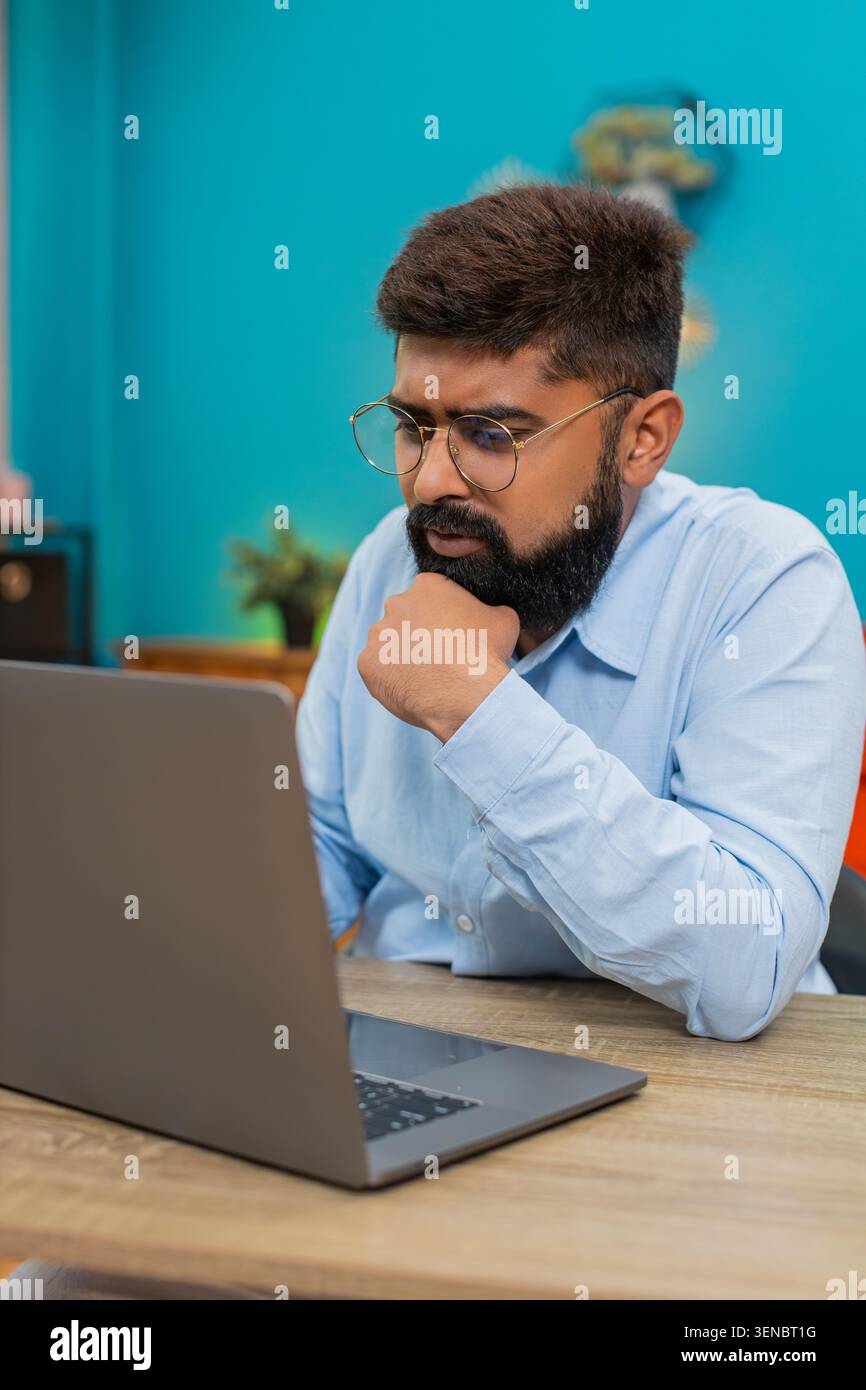 Indian man at home types on laptop stops rubs chin thinking over vital idea creative future plan. Arabian guy at table gazes screen focused eyes show deep concentration solving problem quickly today. Stock Photo