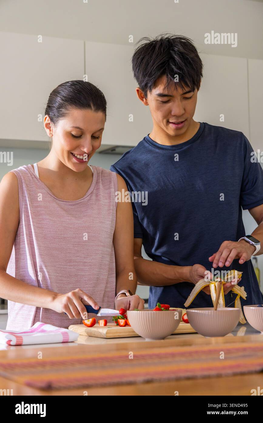 Diverse couple slicing strawberries and peeling banana at counter with beige bowls on wooden board Stock Photo