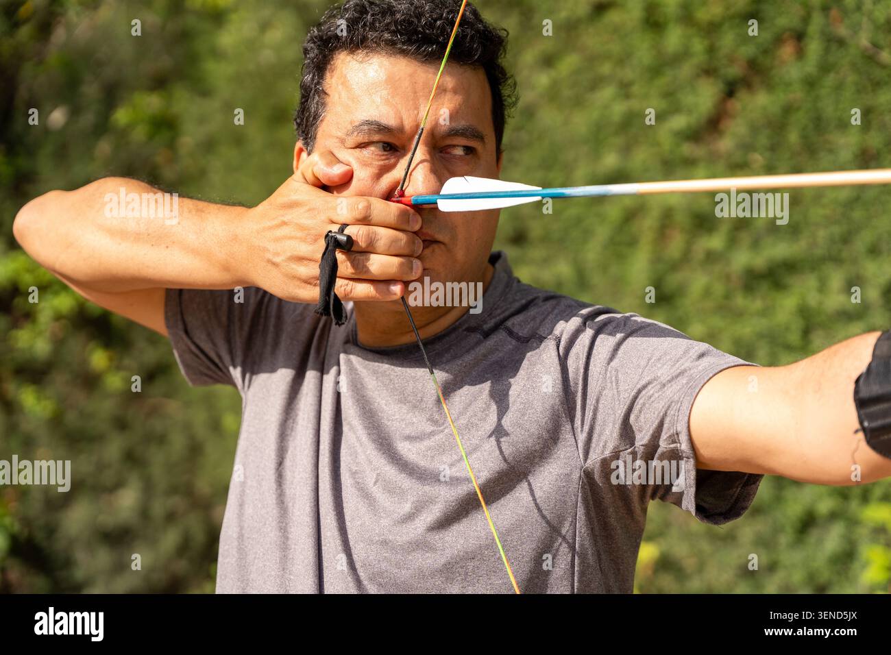Archer concentrating, drawing bowstring with arrow, practicing sport in a green outdoor setting Stock Photo