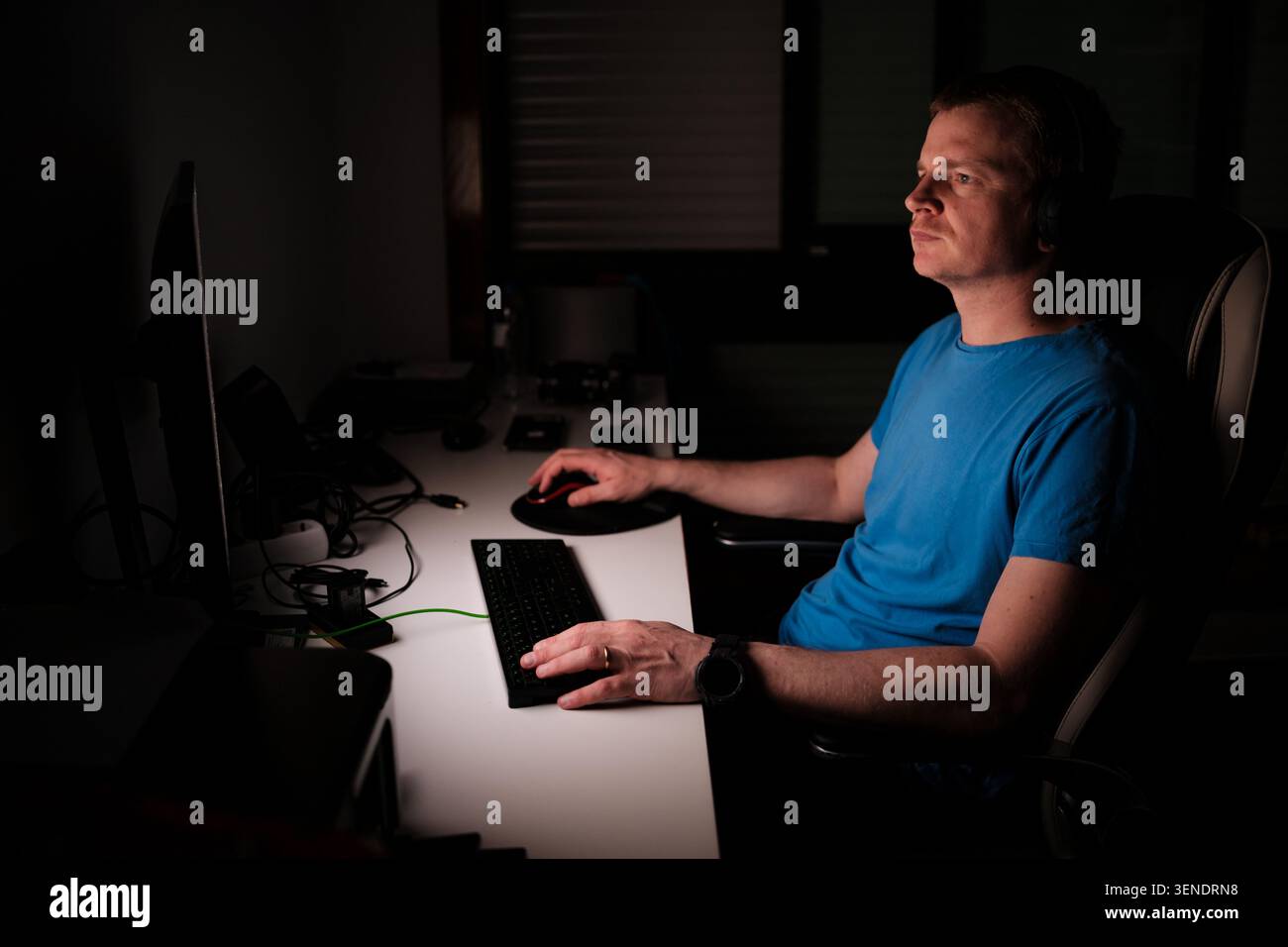 Man concentrating while using a computer at a desk in a dark room Stock Photo