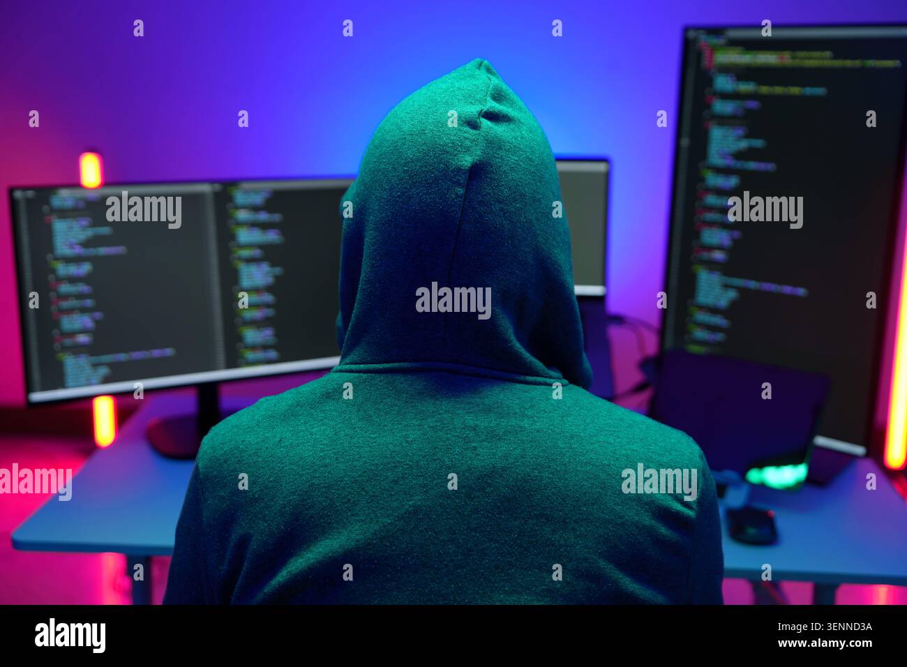 Hooded hacker coding at dual monitors in neon lit room Stock Photo