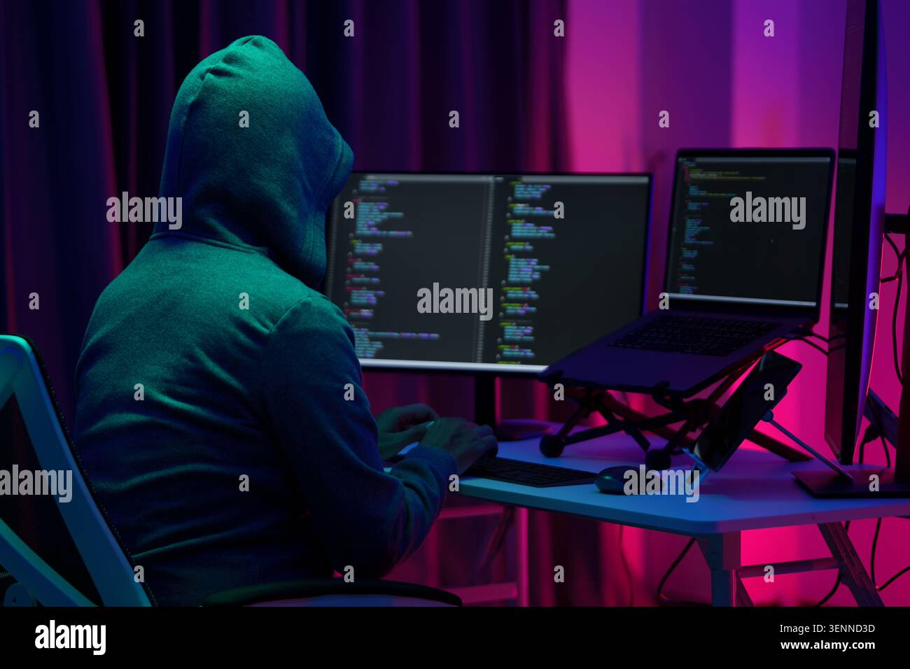 Hooded Hacker Coding at Dual Monitors in Dark Neon Room Stock Photo