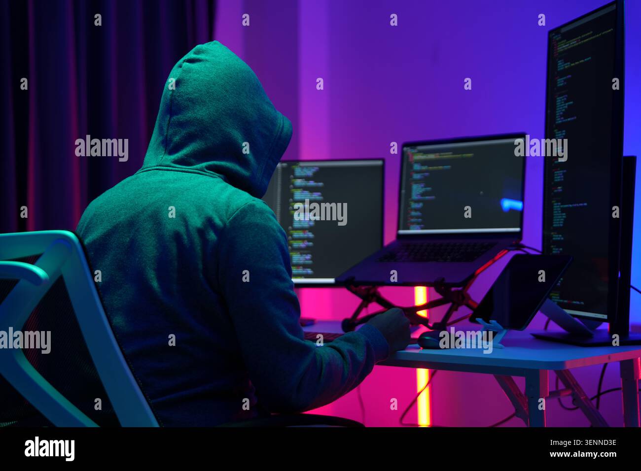 Hooded Hacker Working at Multiple Monitors in Dark Room Stock Photo