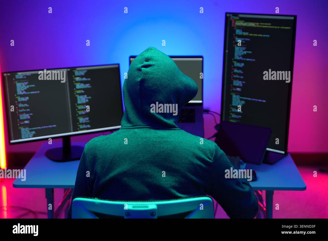 Hacker in Hoodie Working at Multiple Monitors in Neon Light Stock Photo