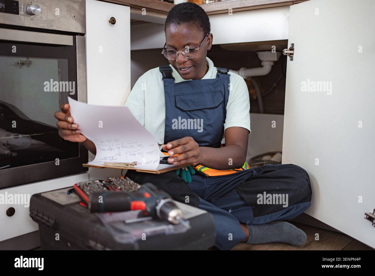 Female plumber checking notes while sitting on kitchen floor Stock Photo