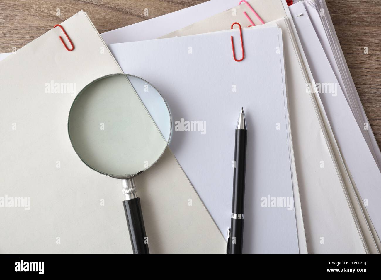 Close-up of a magnifying glass resting on stacked documents and blank papers on a desk, symbolizing inspection, review, research, and office organizat Stock Photo