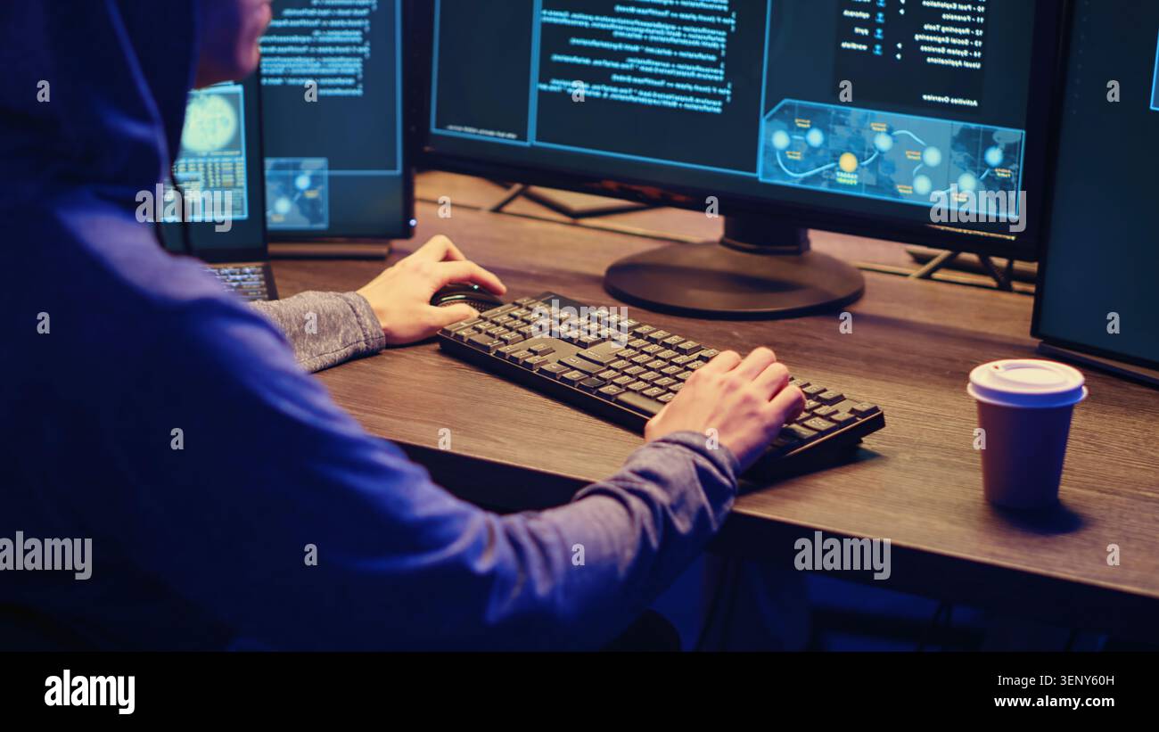 Young hacker drinking coffee while running spyware software designed to gather information from users computers without their knowledge, script kiddie doing illegal stuff in graffiti painted hideout. Stock Photo