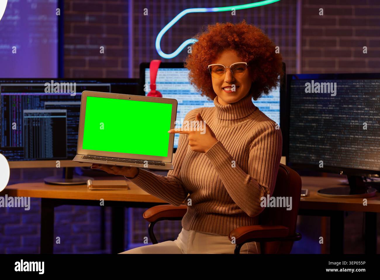 Smiling female developer presenting laptop with green screen displaying software code in dark office, software code is visible on multiple computer mo Stock Photo