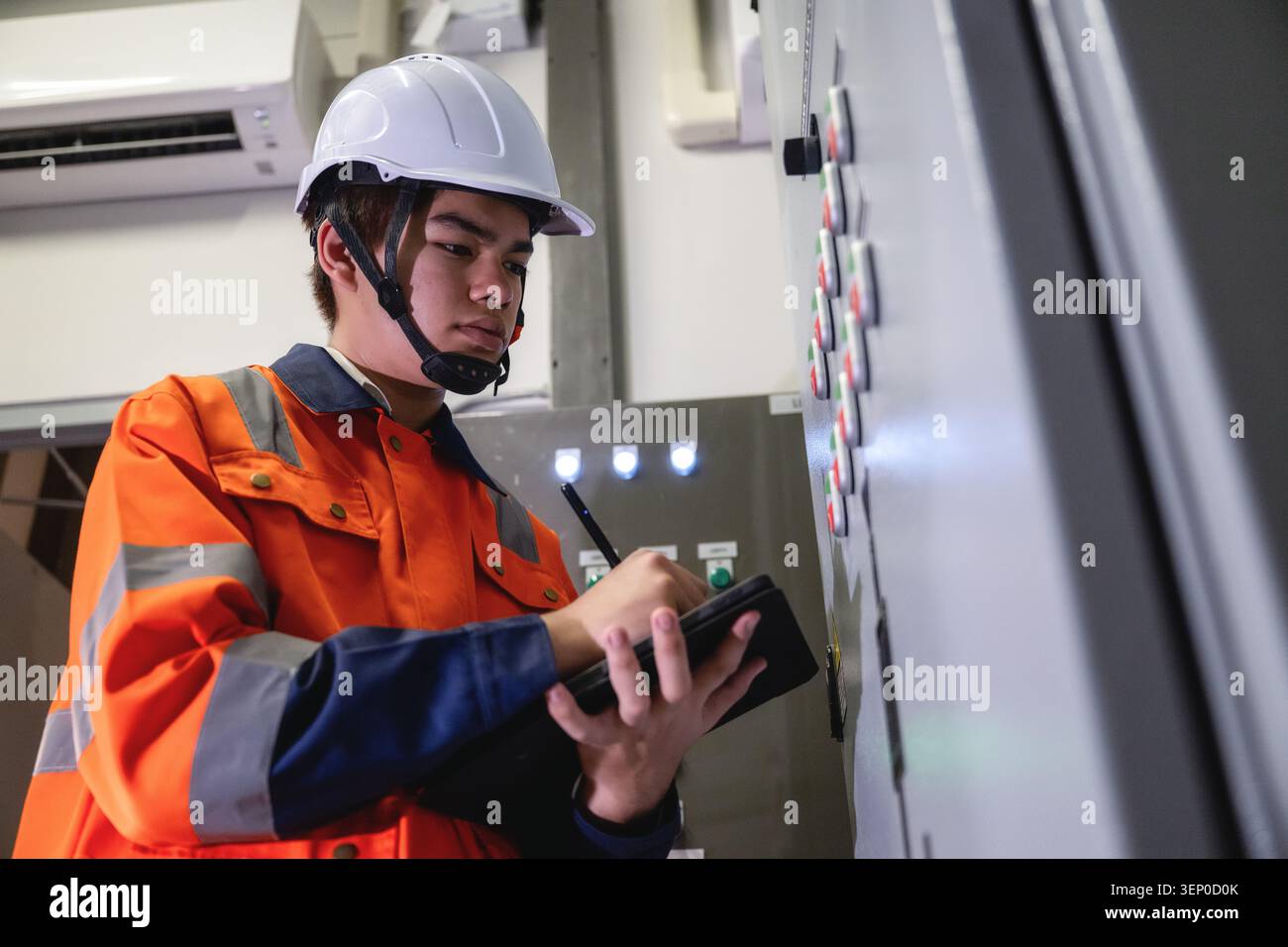 Asian young electrical engineer holding digital tablet inspecting main control panel board and writing data, Inspecting building switch panel factory Stock Photo