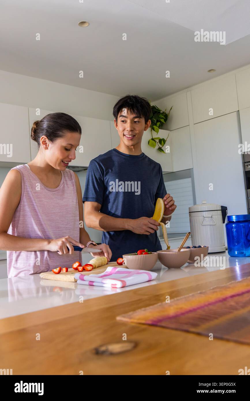 Diverse couple preparing fruit bowls at kitchen island, slicing strawberries and peeling banana Stock Photo