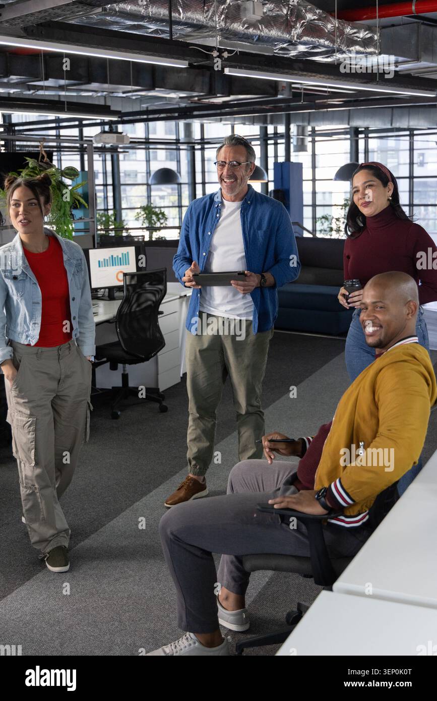 Diverse coworkers talking around senior man holding tablet in open-plan office, denim and mustard Stock Photo