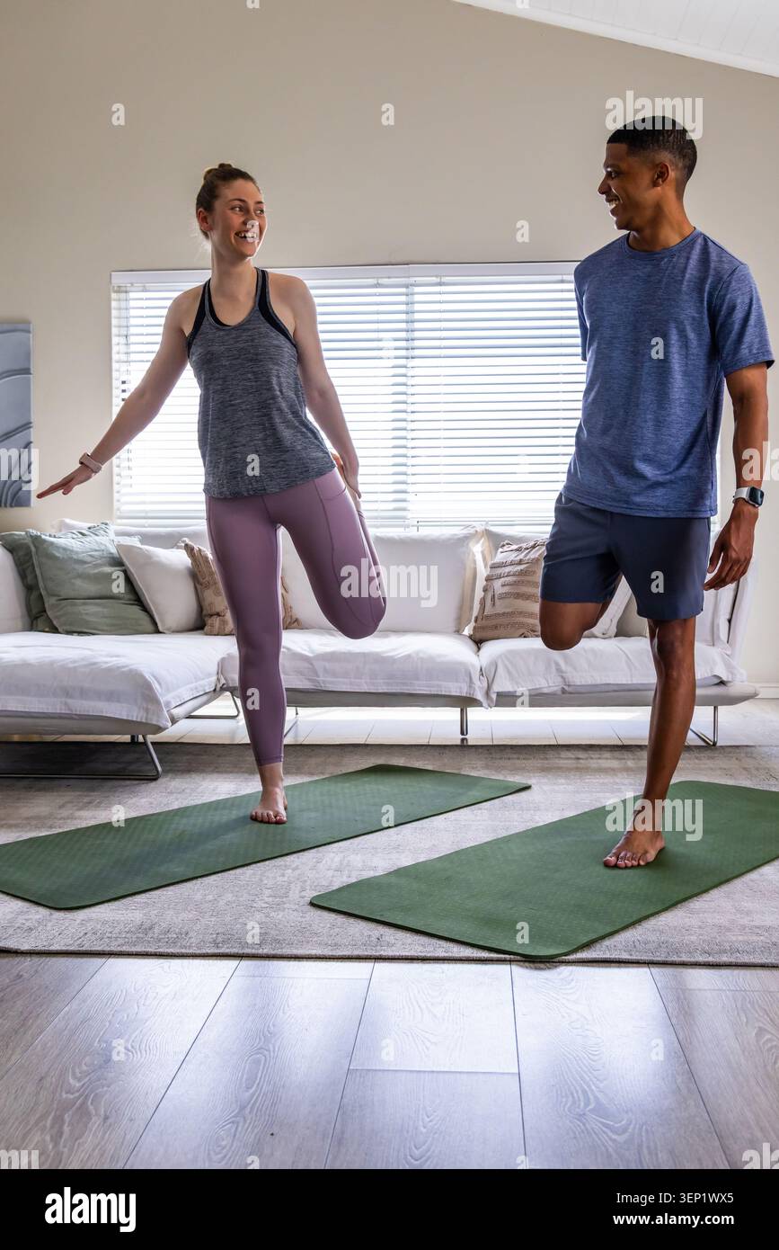 Diverse couple starting workout, doing quad stretch on mats at home wearing sportswear and trackers Stock Photo