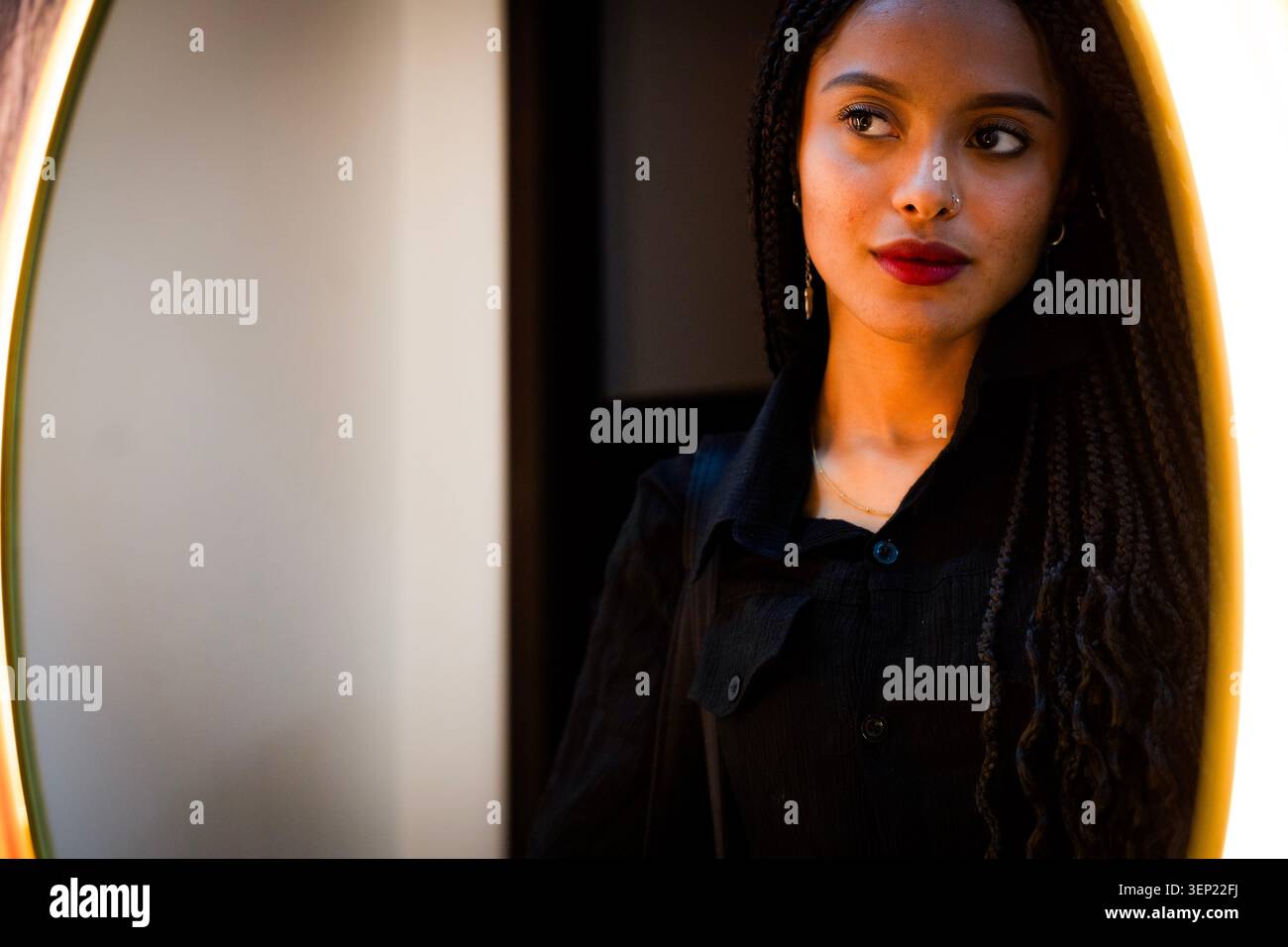 Young mixed race woman looking away in a bright reflection, highlighting her braided hair, red lips, and focused expression, exploring themes of beaut Stock Photo