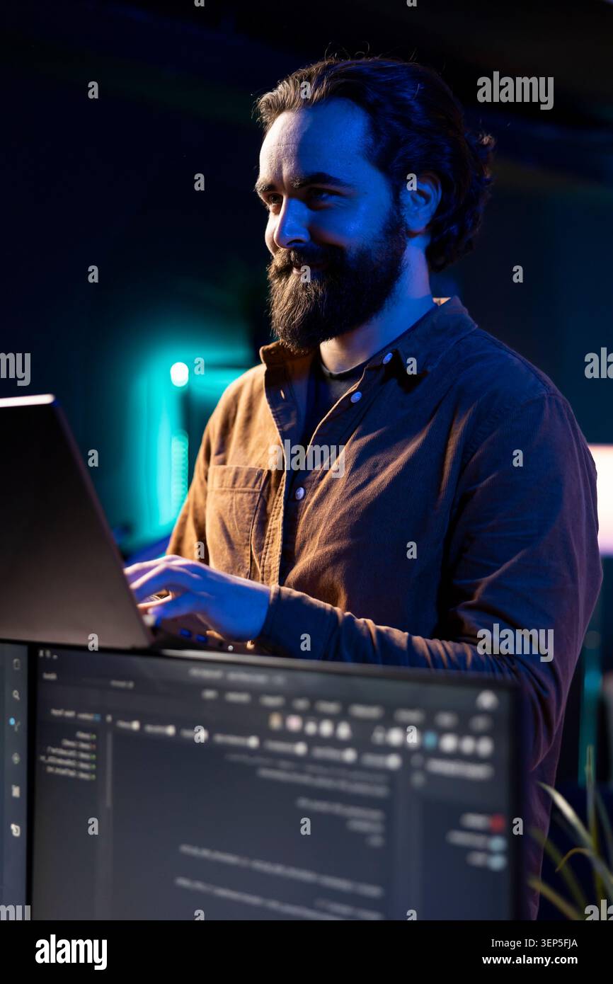 Joyful freelancer doing software debugging using laptop, checking errors in lines of code displayed on PC monitors, cheerful IT professional walking in apartment fixing coding issues using notebook. Stock Photo