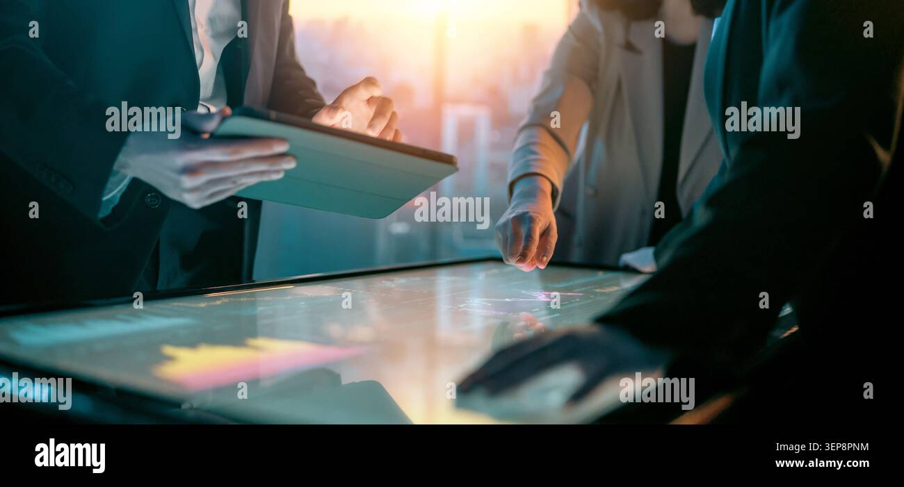 Dynamic image capturing a team of professionals collaborating over a large interactive digital touchscreen table and a tablet, set against a backdrop Stock Photo