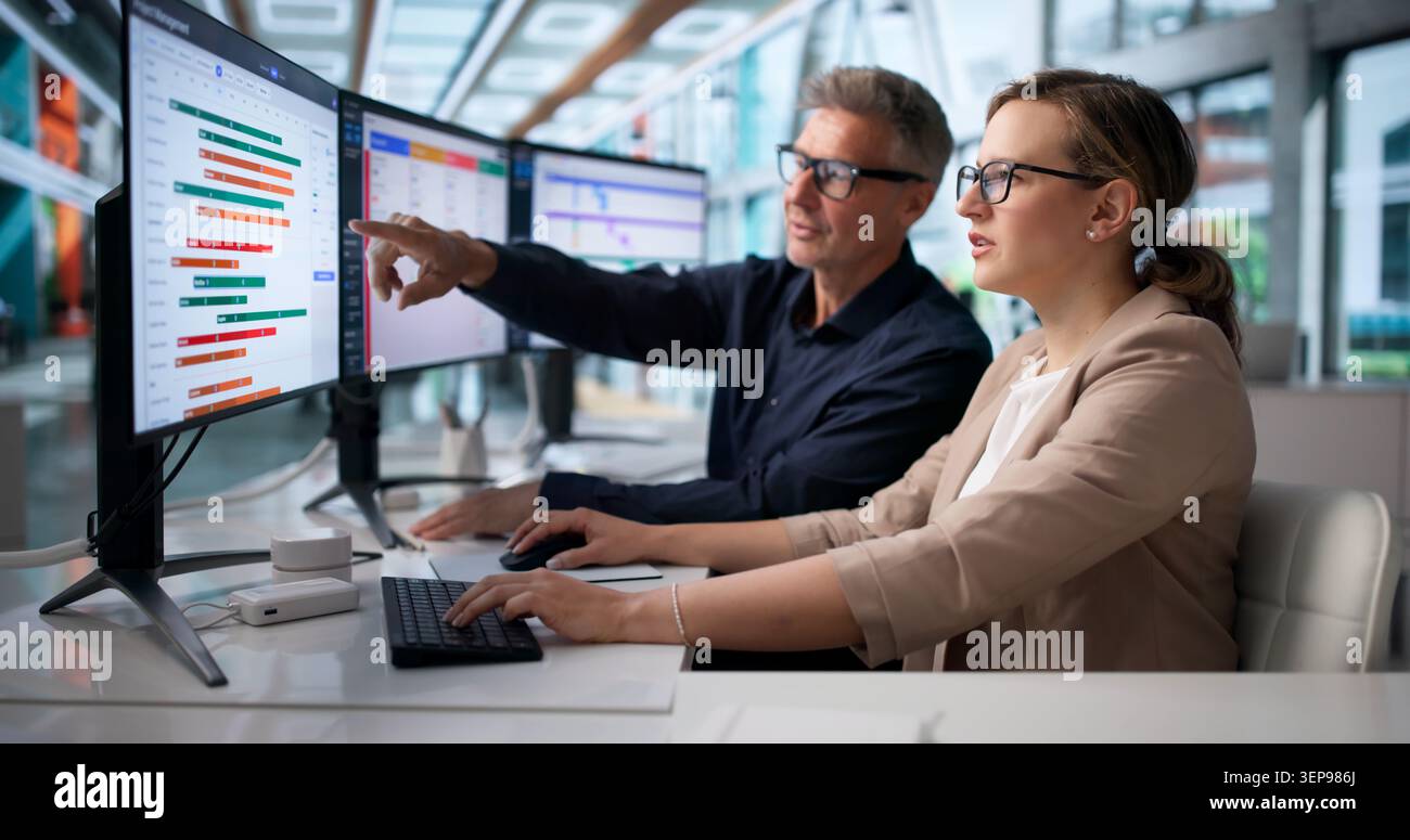 Two Agile Team Members Discuss Project Tasks Using Multiple Computer Screens. Stock Photo