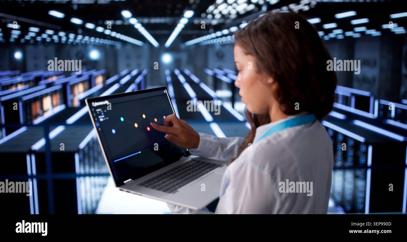 Woman Engineer Monitors Data Servers Using Laptop In Busy Cloud Center. Stock Photo