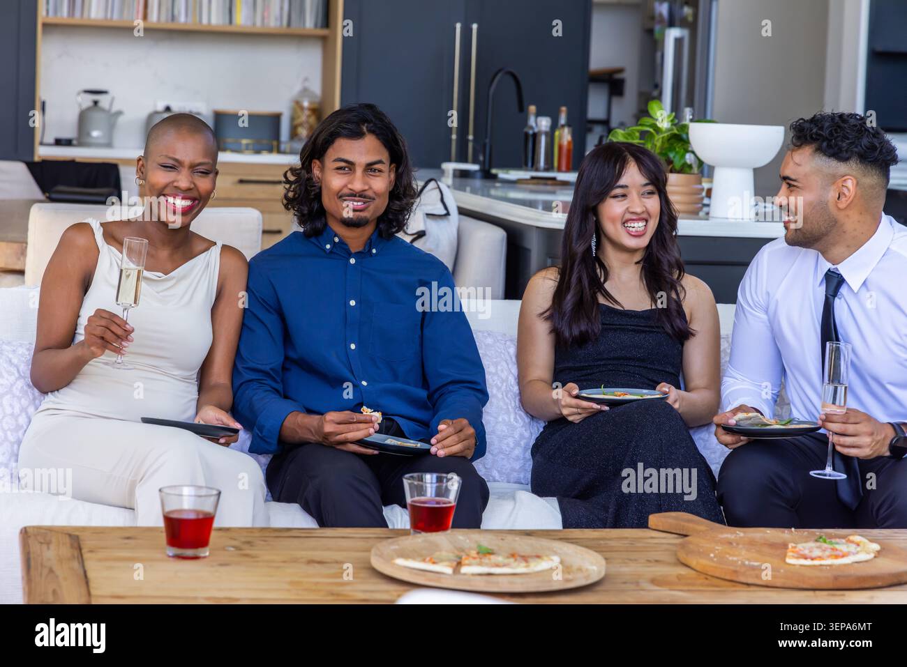 Diverse friends sitting in living room on light sofa sharing pizza and sipping drinks Stock Photo
