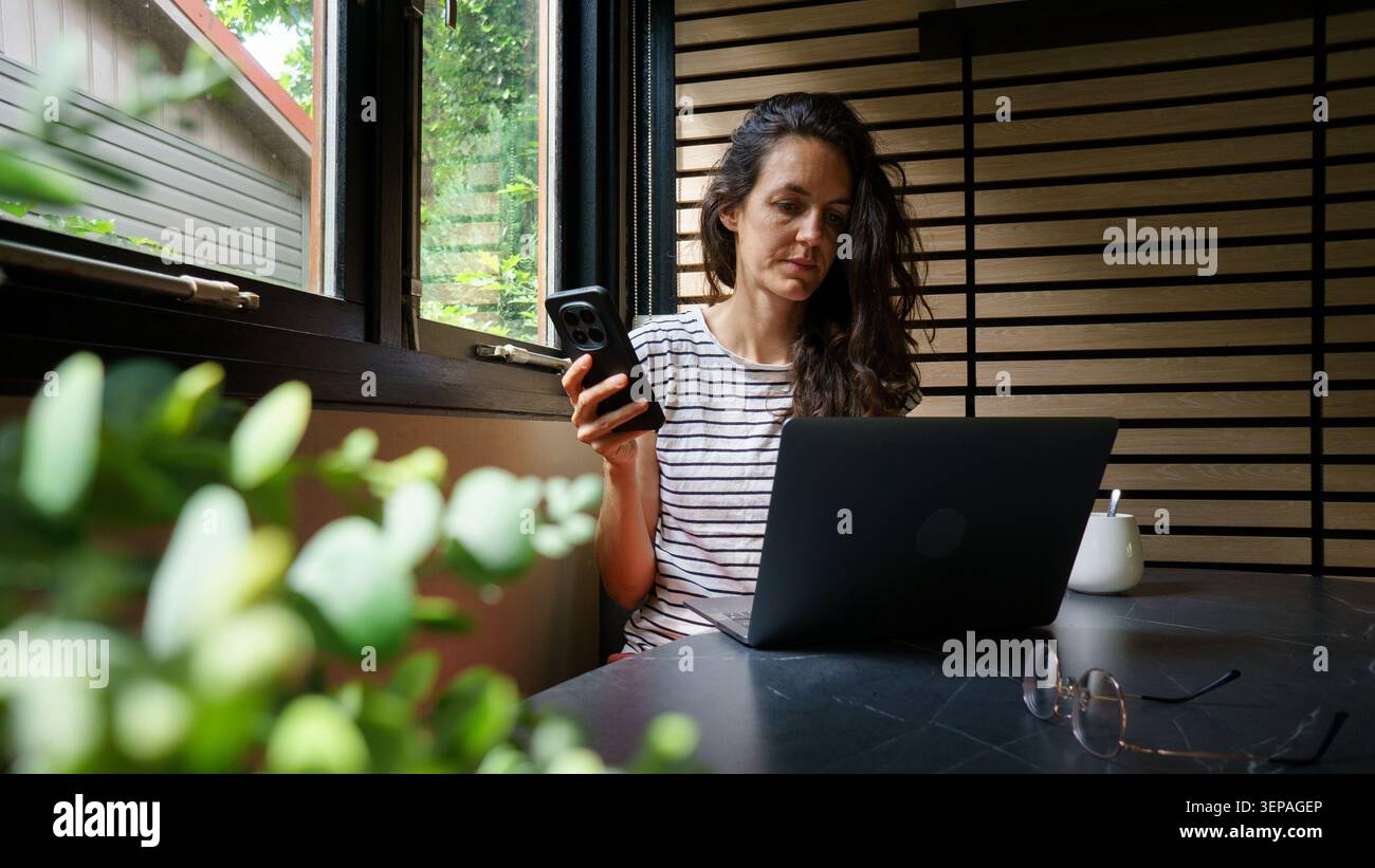 Woman managing multiple communication devices while working at home by a window Stock Photo