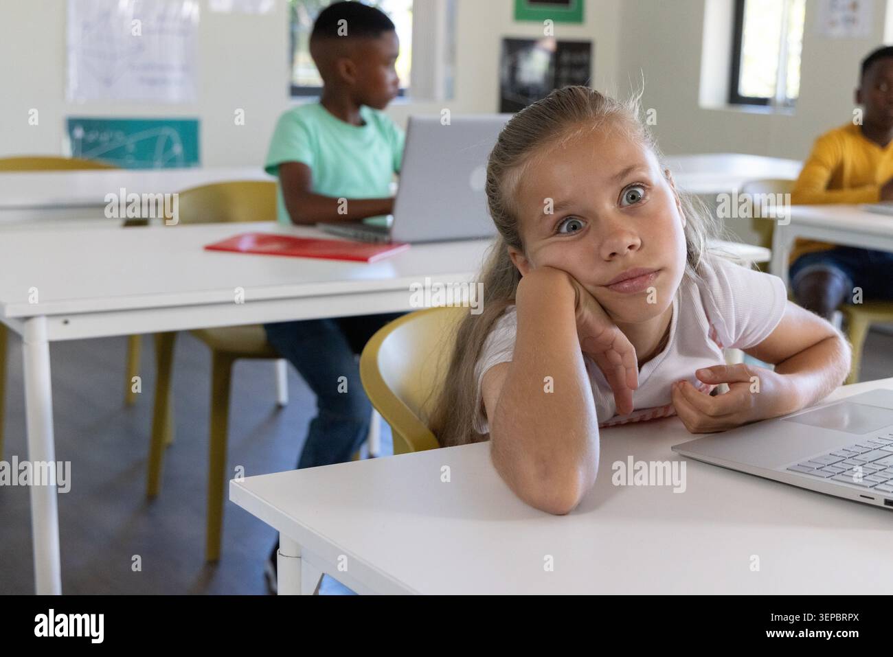 Diverse elementary students sitting at classroom desks using laptops with red notebook and posters Stock Photo