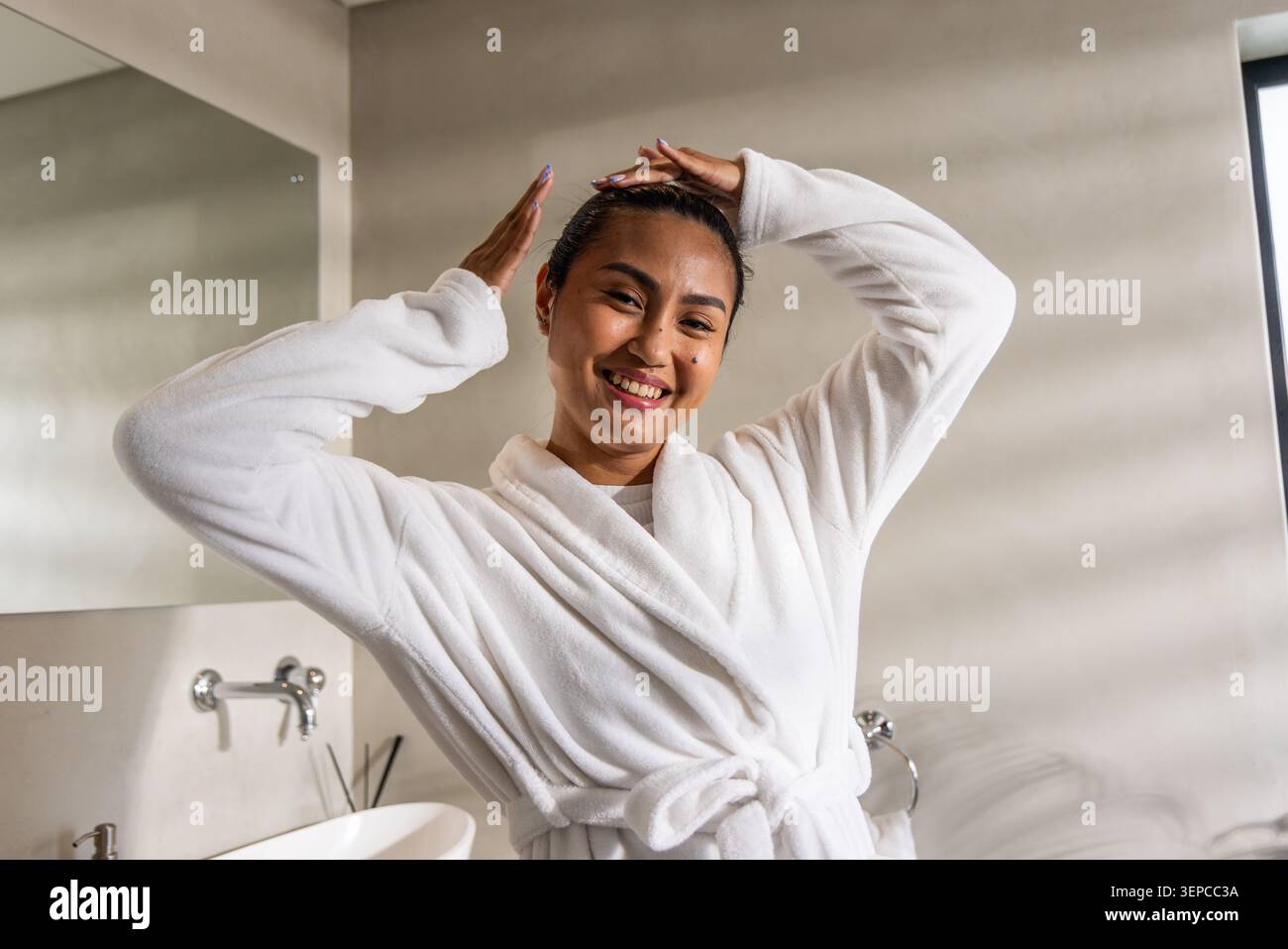 Asian adult woman adjusting hair, smiling in plush white bathrobe in bathroom with chrome faucet Stock Photo