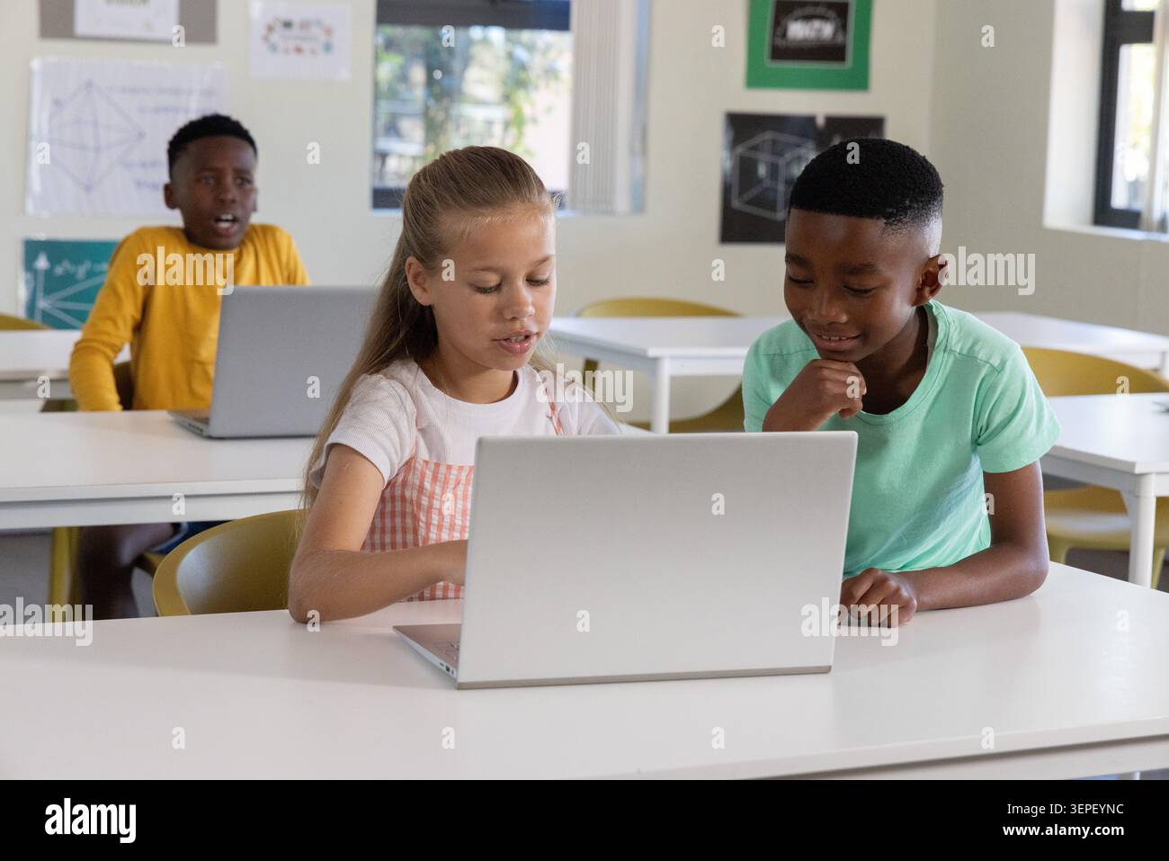 Diverse child classmates collaborating on silver laptop in bright classroom, girl in pink pinafore Stock Photo