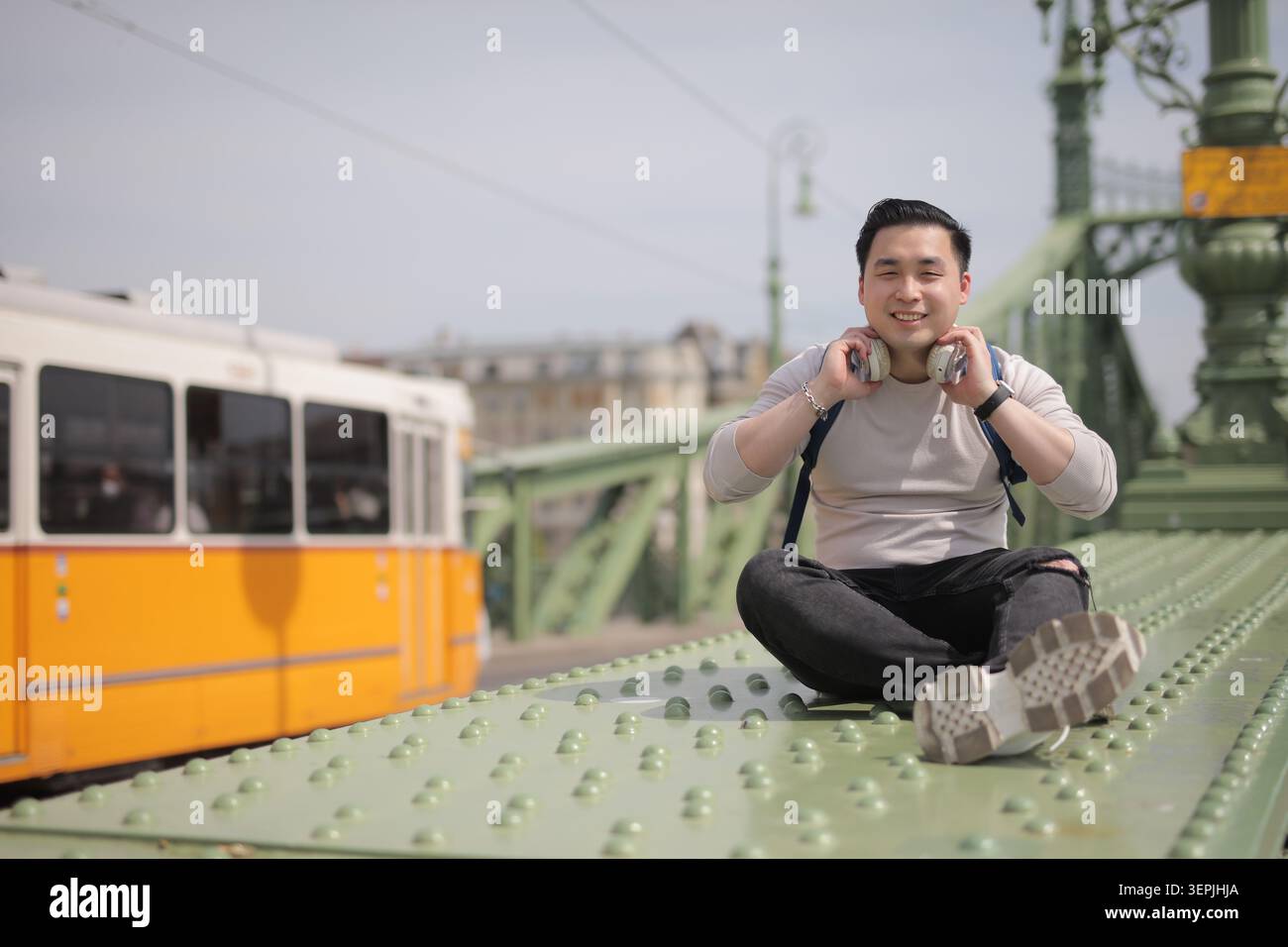 Young Asian man with headphones sits on a city bridge Stock Photo