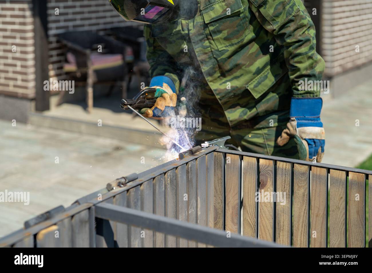 Man in protective welding mask operating welding machine in outdoor residential yard. Performing repair of storage box, home maintenance, safety equip Stock Photo
