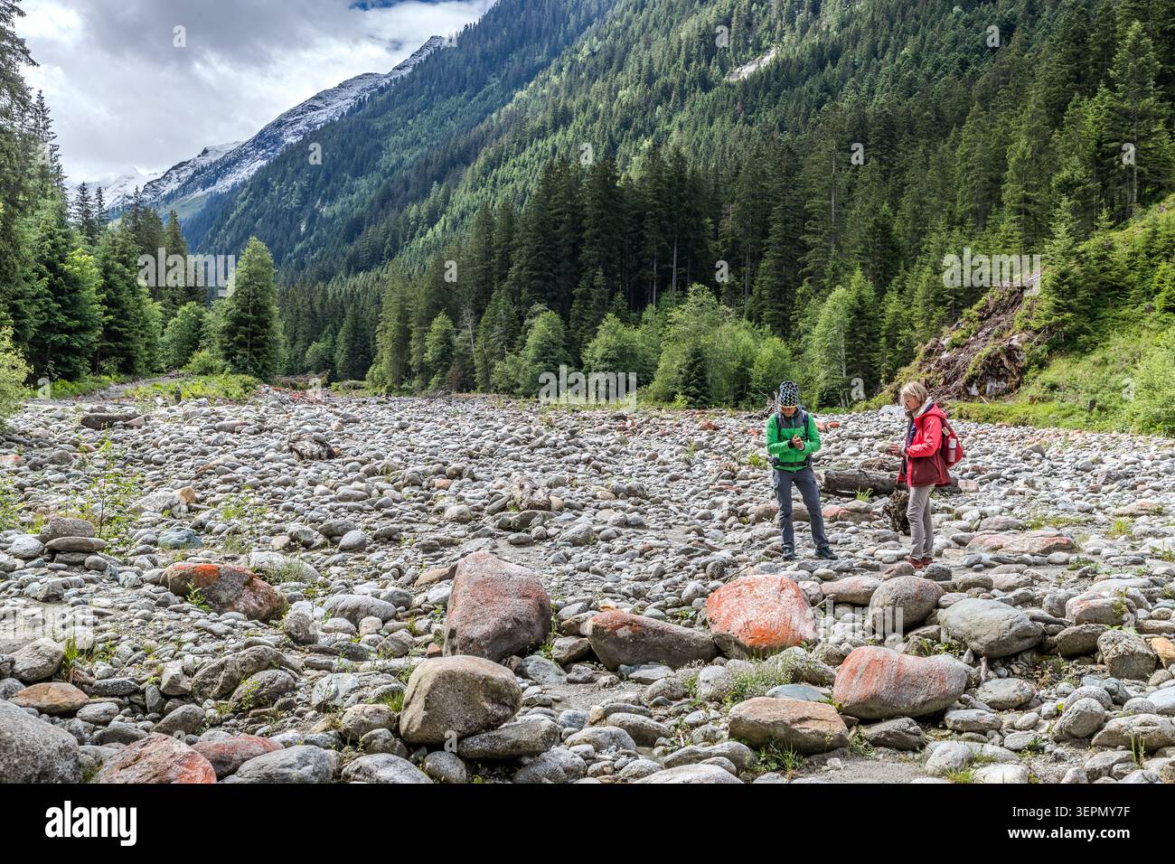 Two hikers are standing in the wide, scree-strewn stream bed of an Alpine valley, with the snow-capped peaks of the Hohe Tauern in the background. Such vast areas of scree are formed by floods and the sediment carried by mountain streams. Sulzau, Rosental, Neukirchen am Großvenediger, Bezirk Zell am See, Salzburg, 5741, Austria Stock Photo