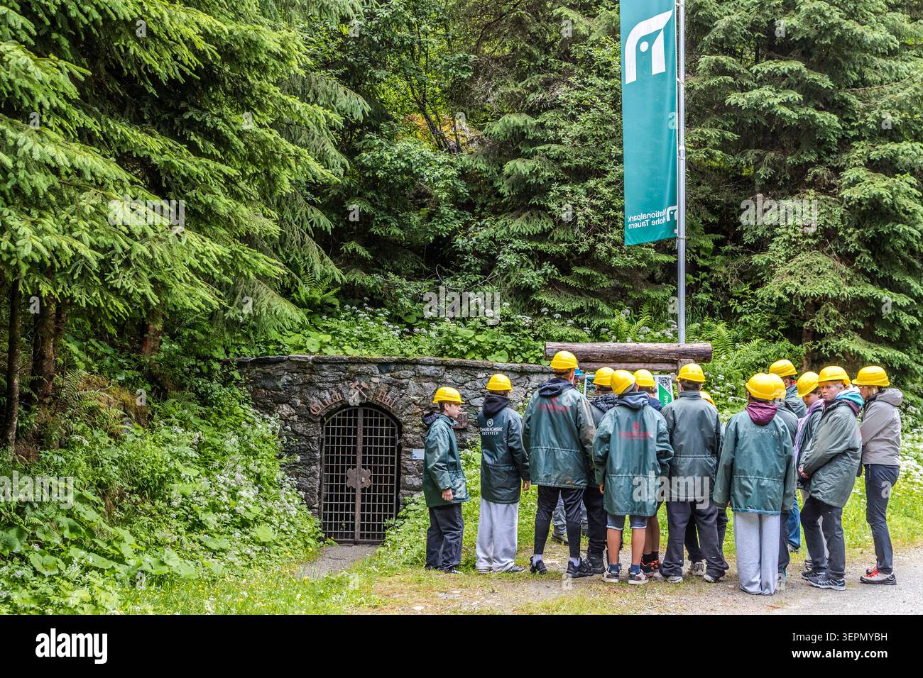 A group wearing yellow safety helmets and green rain capes is standing in front of the brick-built tunnel entrance to the Hochfeld exhibition mine in the Hohe Tauern National Park. The disused tunnel is now used for guided tours exploring the history of Alpine mining. Rosental, Neukirchen am Großvenediger, Bezirk Zell am See, Salzburg, 5741, Austria Stock Photo