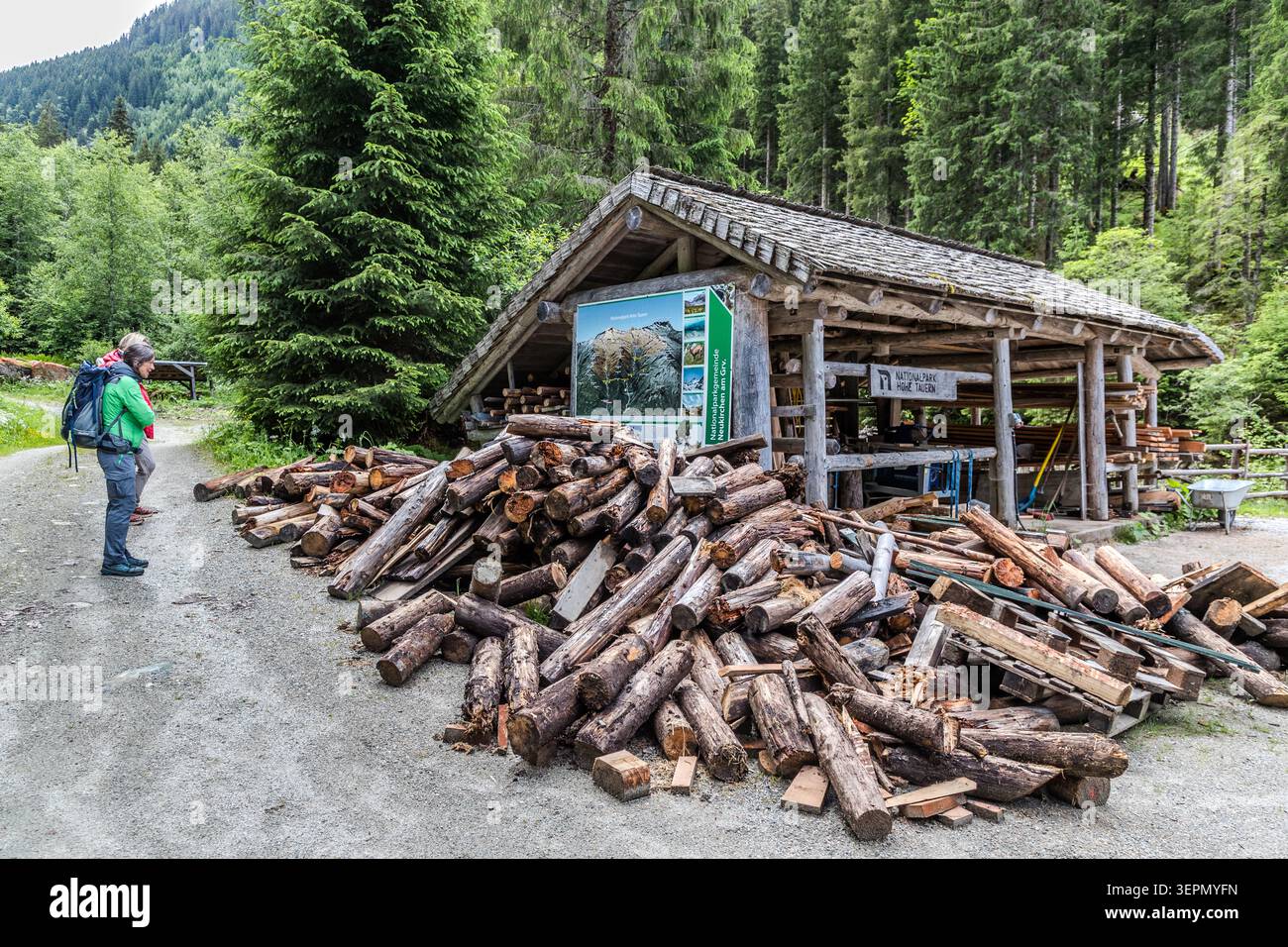 Walker stand in front of a shelter with a National Park information board and a large pile of firewood on the Untersulzbach Waterfall Circular Trail. Shelters like this serve as rest areas and provide shelter from the weather in the Hohe Tauern National Park. Rosental, Neukirchen am Großvenediger, Bezirk Zell am See, Salzburg, 5741, Austria Stock Photo